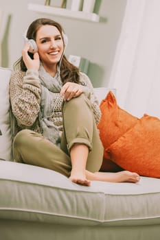 woman with long brown hair sits comfortably on a sofa, smiling as she listens to music through large headphones. Dressed in a cozy sweater and scarf, her relaxed posture adds to the warm, inviting atm