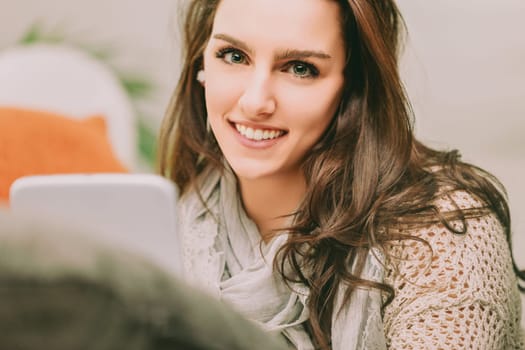 woman with long brown hair smiles brightly while using a digital tablet for online shopping. Dressed in a cozy sweater and scarf, she exudes warmth and happiness in a comfortable, modern setting
