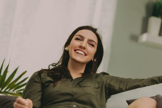 Woman with dark, straight hair smiles broadly while lounging on a modern, light sofa. Dressed casually in a green top and white shorts, her expression radiates warmth and happiness