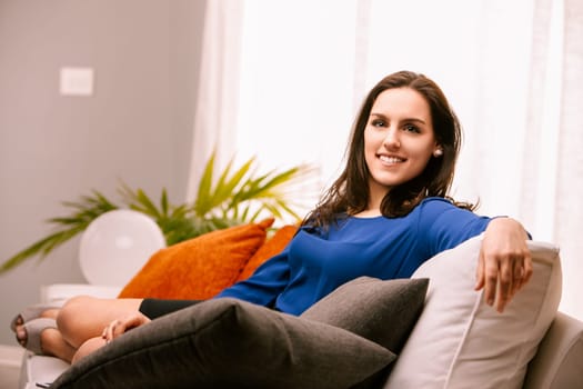 Young woman smiles on her white sofa, enjoying a relaxing day at home in her modern apartment, surrounded by cushions and pillows in a peaceful, happy atmosphere filled with natural light