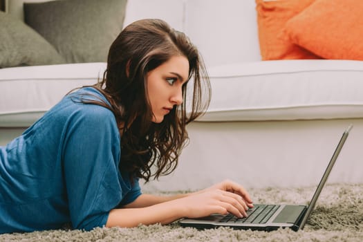 woman with long brown hair lies on the carpet, intently focused on her laptop. Dressed in a blue top, her posture suggests deep concentration as she types, immersed in her work