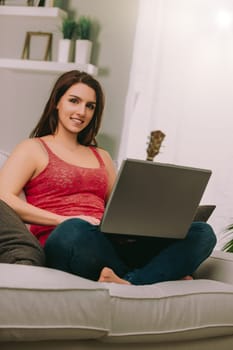 young woman with a bright smile and brown hair sits on a sofa with a laptop on her lap. Dressed casually in a pink lace top and jeans, she exudes warmth and approachability