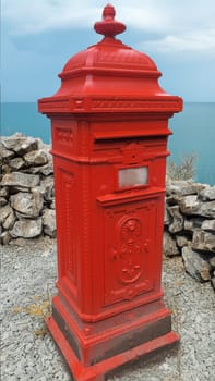 Classic, red British mailbox stands alone on a stone wall, with a view of the wide sea