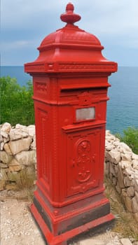 Classic, red British mailbox stands alone on a stone wall, with a view of the wide sea