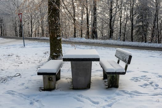 Picnic bench and table in a parking lot in the snow in a forest with a street