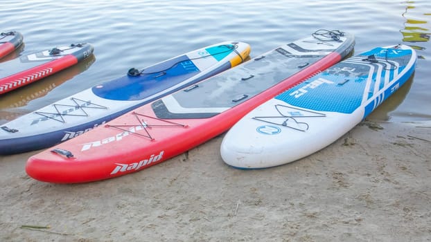 Empty SUP boards sway on the waves on a calm river or lake. Water sports. Lots of boards at the rental shop, the sun reflecting off the ripples of the water. Panning. Ukraine, Kyiv - August 07, 2022