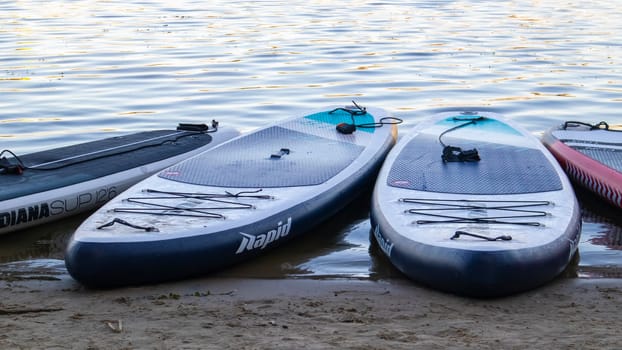 Empty SUP boards sway on the waves on a calm river or lake. Water sports. Lots of boards at the rental shop, the sun reflecting off the ripples of the water. Panning. Ukraine, Kyiv - August 07, 2022
