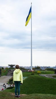 A little girl stands and looks at the flag of Ukraine on a high flagpole. Ukrainians are against the war. Drawing attention to military operations in Ukraine. Ukraine, Kyiv - October 9, 2022