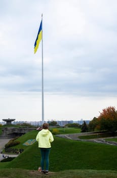 A little girl stands and looks at the flag of Ukraine on a high flagpole. Ukrainians are against the war. Drawing attention to military operations in Ukraine. Ukraine, Kyiv - October 9, 2022