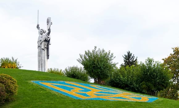Statue of the Motherland against the blue sky. Coat of arms of Ukraine on the lawn, lined with blue and yellow stones. The trident, the official symbol of the state. Ukraine, Kyiv - October 08, 2022