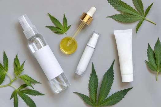 Cosmetic bottles, jars and pipette with blank labels near green cannabis sativa leaves on a grey table top view. Mockup, Copy space. CBD Organic skincare beauty products.