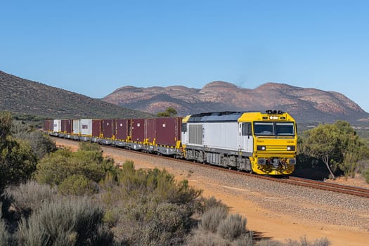 Yellow locomotive pulling cargo containers through dry, mountainous terrain with scrub vegetation