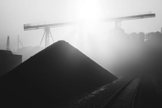 Black and white image of industrial cranes and coal piles shrouded in fog, creating a dramatic atmosphere.