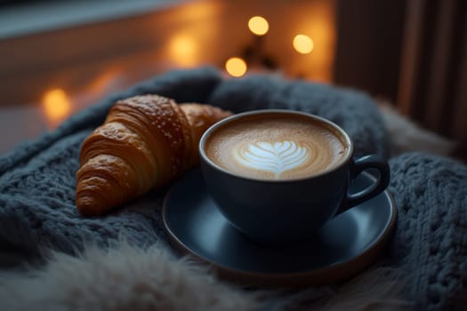 Dark cup of latte with leaf design in foam, croissant on saucer, soft knit fabric, fairy lights in background.