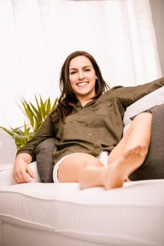 Young woman is sitting on her couch at home, enjoying a relaxing day. She is wearing casual clothing and has a happy expression on her face