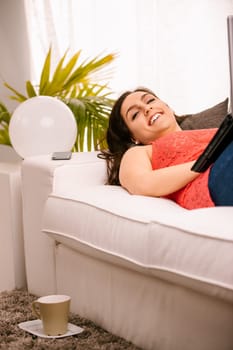 Young woman happily browsing the internet on her laptop while lying on her cozy sofa in a bright living room
