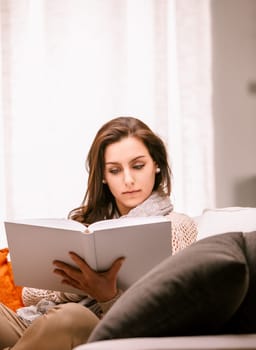 Young woman relaxes on her cozy couch at home, immersed in a captivating book, enjoying a peaceful moment of tranquility
