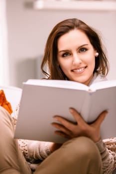 Young woman enjoying a peaceful moment at home, curled up on the sofa with a captivating book, fully immersed in reading and beaming with contentment