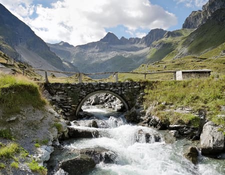 White campervan with red accents, stands on a small bridge over a roaring mountain stream. An impressive mountain backdrop rises in the background.