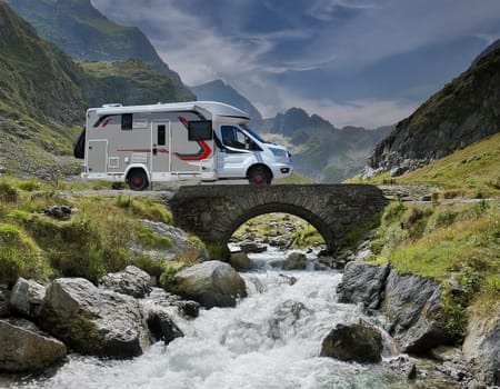 White campervan with red accents, stands on a small bridge over a roaring mountain stream. An impressive mountain backdrop rises in the background.