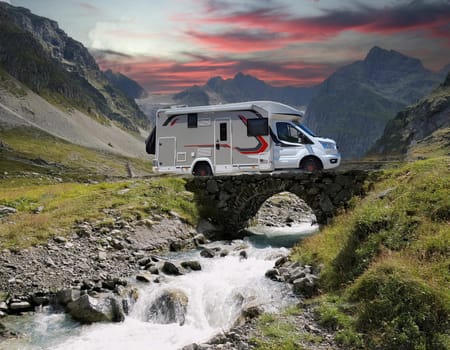 White campervan with red accents, stands on a small bridge over a roaring mountain stream. An impressive mountain backdrop rises in the background.