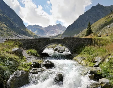 White campervan with red accents, stands on a small bridge over a roaring mountain stream. An impressive mountain backdrop rises in the background.