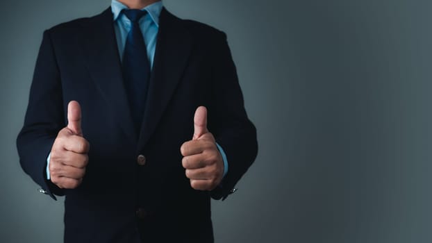 A businessman in a suit stands with his arms crossed and thumbs up on a dark background.