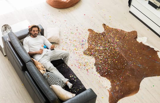 Man lying on black leather couch and holding microphone in disheveled room with colorful confetti on wooden floor after Christmas celebration party at home.