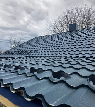 Close-up house roof with steel roofing sheet. Traditional metal shingle tiles. Blue modular tile effect roof panel. Profiled metallic corrugated pantile as clay or concrete slate. Building's covering