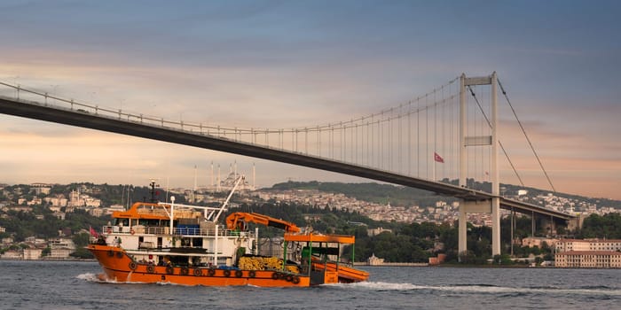 Istanbul, Turkey - August 8 2022: A workboat navigates the waters beneath the Bosphorus Bridge, showcasing the vibrant colors of sunset over the city