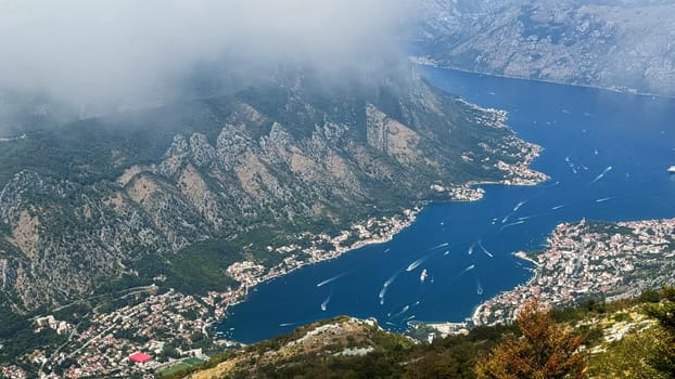 Aerial view of a coastal town and deep blue bay nestled among rugged mountains with distinct rock formations. Fog and clouds
