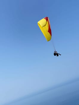 People flying on a paraglider with a yellow and red parachute high in the sky, surrounded by a vast expanse of clear blue. extreme sports, outdoor activities and aerial photography stocks.