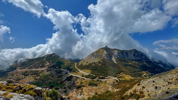 A breathtaking view of a mountain range with a winding road, surrounded by lush greenery and dramatic cloud formations against a bright blue sky.suitable for travel promotions and wallpapers.