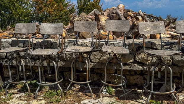 A close-up of six wooden bar stools with rusty metal frames, lined up against a stone wall, against a background of chopped wood and green trees under a clear sky.decay and outdoor decor themes.