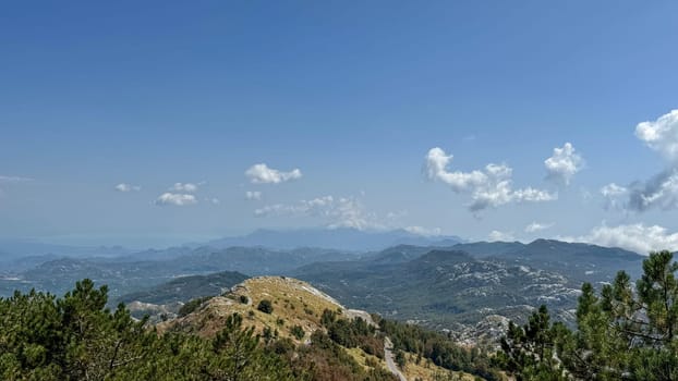 Wide mountain view with distant peaks and hills under clear, bright blue sky. The foreground is dotted with trees highlighting the natural scenery.