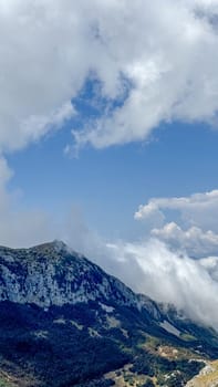 A dramatic scene of a mountain peak covered in dense greenery, partially hidden by swirling clouds under a bright blue sky.