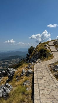 A scenic stone-paved path winding up a grassy mountain slope, surrounded by rocks and leading towards a horizon filled with mountains and clear blue skies.