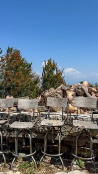 Old weathered wooden high bar stools with rusty metal frames against a background of green trees, piles of firewood and clear blue sky.