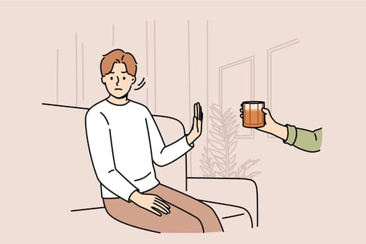 Determined man refuses alcohol, demonstrates stop gesture, sitting on sofa near hand with whiskey. Guy says no to alcohol and wants to live sober lifestyle to avoid hangover and intoxication