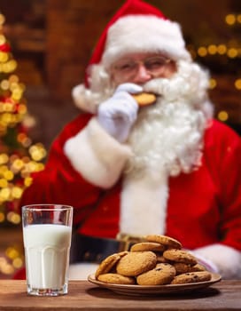 Cookies and milk mug on the table with Santa Claus in the background eats the tasty biscuit next to Christmas tree. Wonderful winter holidays scene