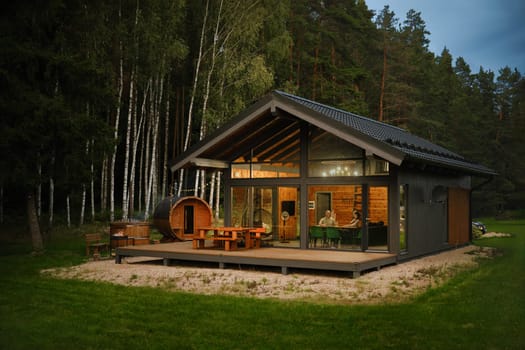A modern cabin with a large glass windows and wooden deck with patio furniture at a forest clearing and a barrel-shaped wooden sauna