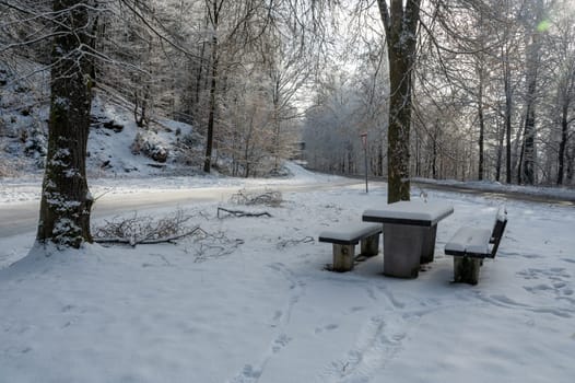 Picnic bench and table in a parking lot in the snow in a forest  with a street