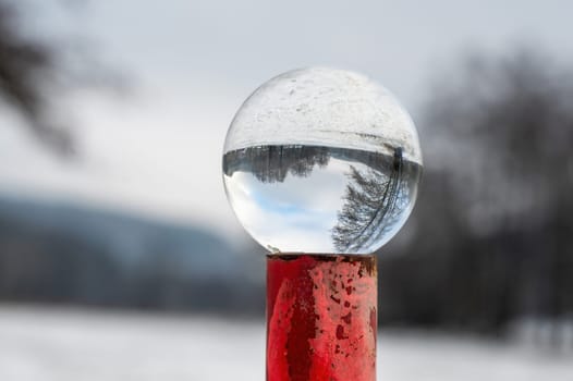 Glass ball lying on a rusty red metal post on a cold winter day in nature, with reflections of a snowy landscape in the glass