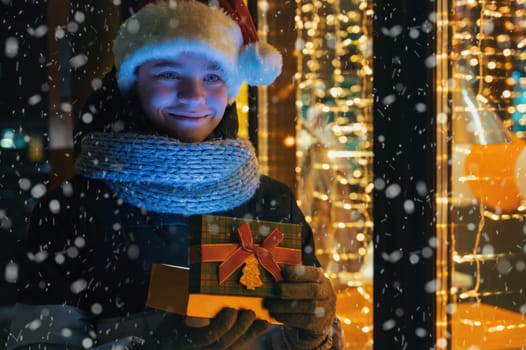 Boy in Santas hat with gifts box near illuminated shop window. Xmas presents holidays, or shopping on New Year or Christmas concept