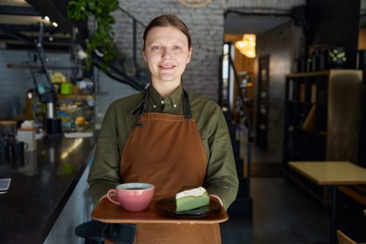 Woman barista giving wooden tray with freshly brewed coffee drink in ceramic cup and cake piece portion on plate. Client service at cafeteria or restaurant