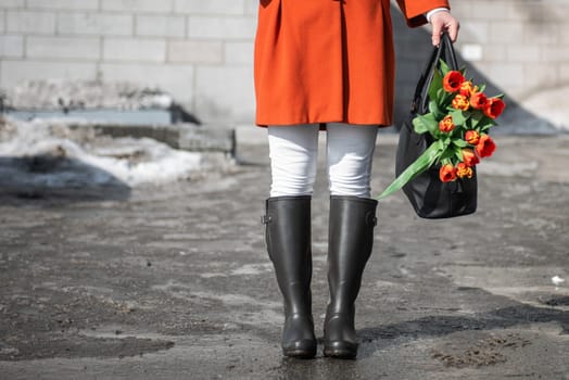 Closeup of woman in a orange coat and rubber boots walking down the street with a bouquet of tulips in spring city