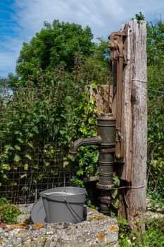 A rusty old water pump is sitting next to a bucket. The bucket is filled with water and is placed on the ground. The scene has a rustic and somewhat abandoned feel to it