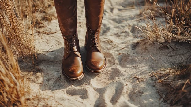 Close-up of brown leather boots standing on sand with dry grass, capturing a sense of adventure and outdoor exploration.