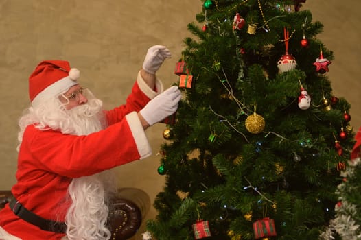Happy Santa Claus in red suit decorating Christmas tree with balls, gift boxes and festive figurines in living room at home. Christmas and New Year concept.