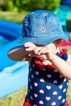 A little girl in a panama hat covered her eyes with her hands outdoors near an inflatable pool